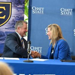 A man in a suit shakes the hand of a woman in a suit in front of ETSU and ChattState backdrops.