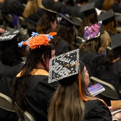 A woman sititng in a crowd of graduates wears a decorated mortaboard that reads "Class of 2025"