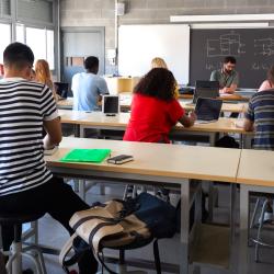 A group of students in a classroom face a teacher and blackboard with electrical diagrams on it.