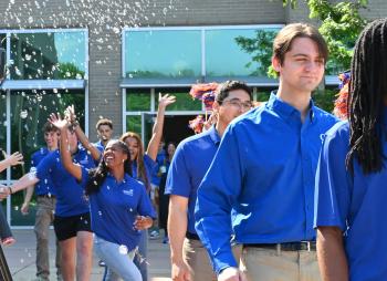 Students in Blue SkillsUSA shirts queue to board a bus to Atlanta. In the background, two young women reach for bubbles in the air as they celebrate. Students in Blue SkillsUSA shirts.