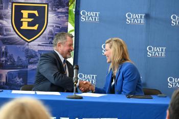 A man in a suit shakes the hand of a woman in a suit in front of ETSU and ChattState backdrops.