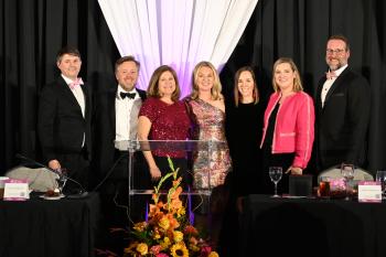 Eight people in formal wear stand in spotlights on a stage during Dinner of Firsts 2026.