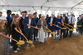 A line of people in hard hats toss dirt with ceremonial shovels.